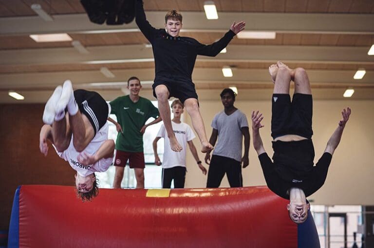 Flere unge mennesker udfører akrobatiske spring på en trampolin. Nogle hopper op i luften, mens andre er i gang med at lave baglæns saltoer. Atmosfæren er fyldt med energi og entusiasme, og der er en tydelig følelse af teamwork og sjov blandt deltagerne.