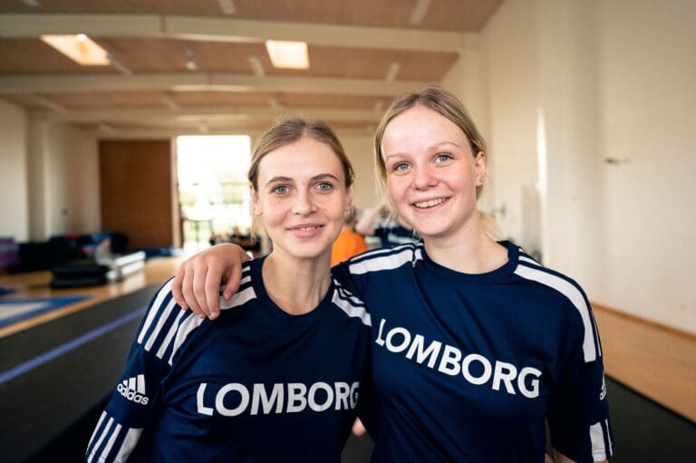 To unge kvinder står tæt sammen med smil på læberne. De bærer matchende trøjer med teksten 'LOMBORG'. Baggrunden viser en lys træningshal med udstyr til gymnastik og sport. Atmosfæren er venlig og energisk, hvilket indikerer en aktiv træningssession.