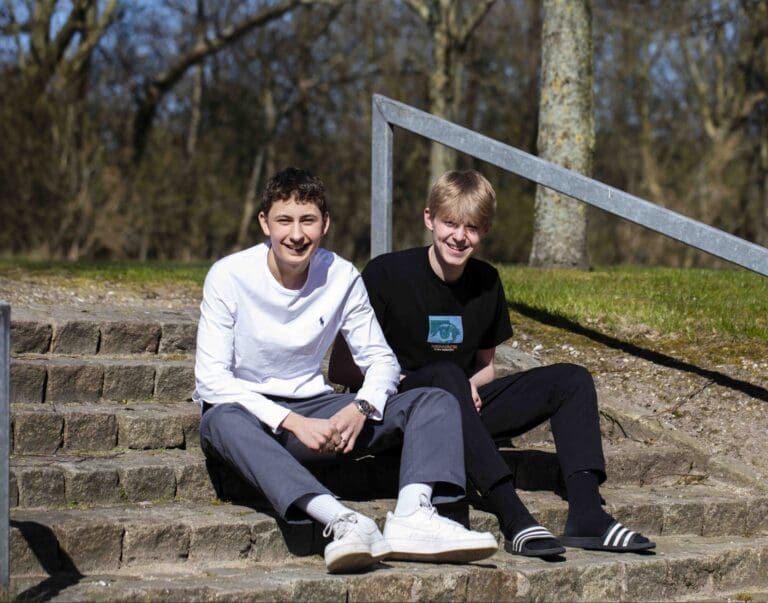 To unge mænd sidder på en trappe i et solrigt miljø. Den ene har en hvid trøje på, mens den anden bærer en sort t-shirt. Begge ser glade ud og nyder det gode vejr.