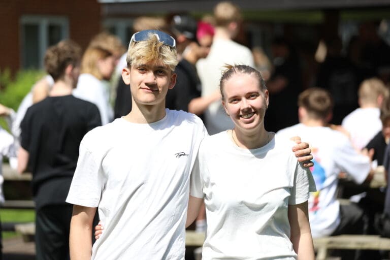 To unge mennesker står tæt sammen og smiler. De bærer begge hvidt tøj og ser glade ud. I baggrunden ses en gruppe mennesker, der socialiserer og nyder det gode vejr.